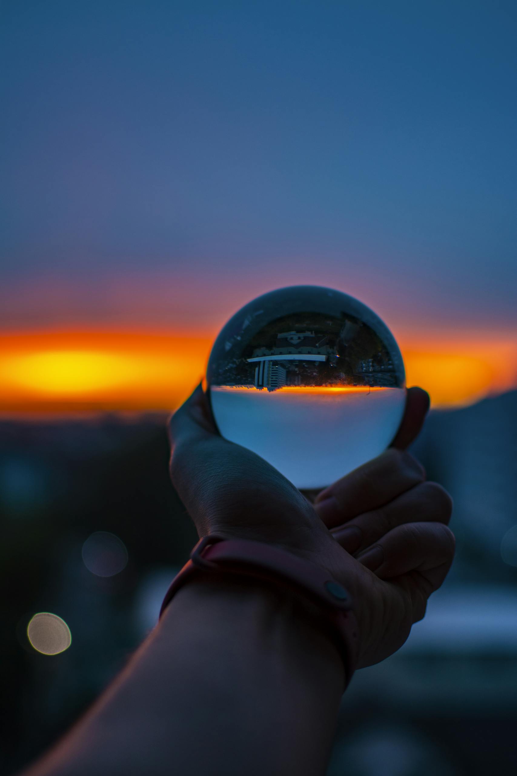 Unrecognizable person with sunset sky reflecting upside down in glass ball in sundown time in city on blurred background outside